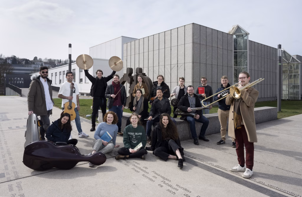 Gruppe junger Musiker:innen mit Instrumenten vor modernem Gebäude, fröhlich und entspannt, im Hintergrund „HFM Saar“.